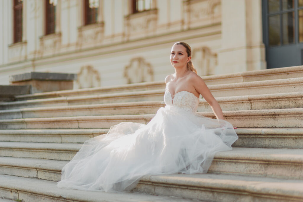 Bride sitting on stairs at the entrance of Belvedere Castle in Vienna, posing in the sunlight during her wedding photoshoot.