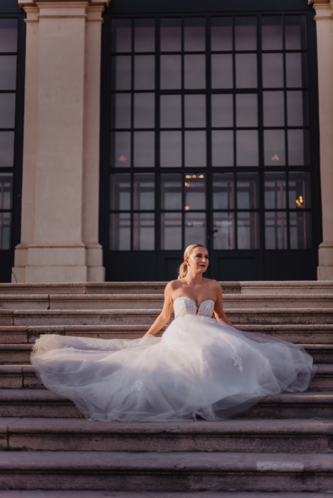 A bride elegantly seated on the grand staircase of Schönbrunn Palace, her flowing gown draping over the steps, framed by golden baroque architecture in soft natural light.