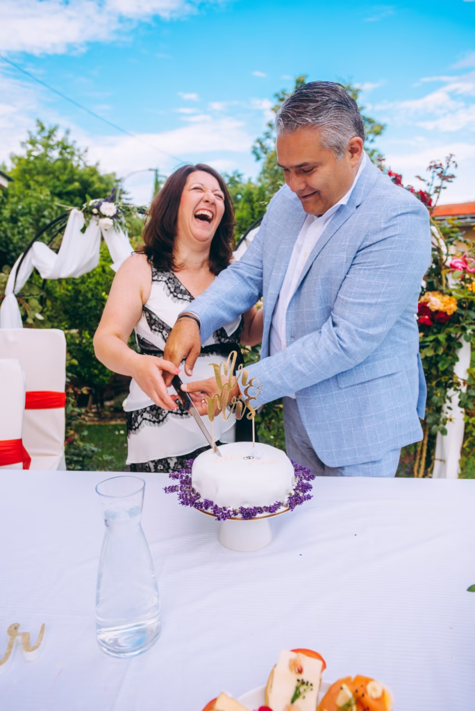 A bride and groom laughing joyfully as they cut their wedding cake, surrounded by an opulent cake table with floral arrangements and soft candlelight, capturing a luxury wedding moment.