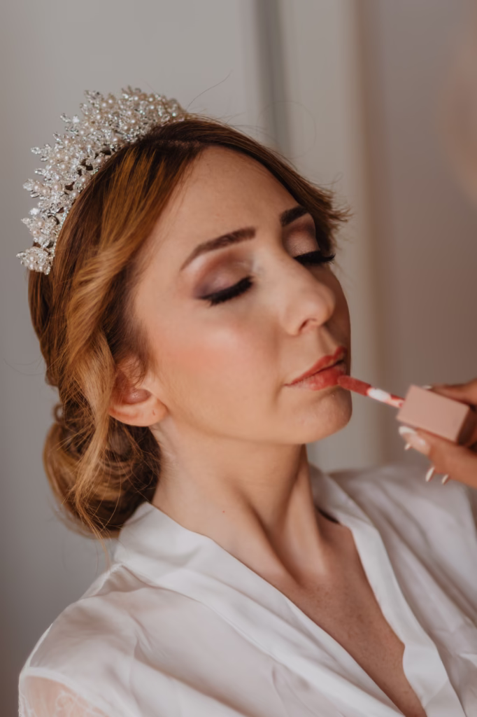 Bride getting ready on her wedding day, adjusting her pearl necklace while wearing an elegant satin gown with a long train. Soft natural light highlights her delicate features as she holds a bouquet of white roses, with a sparkling crown completing her timeless bridal look.