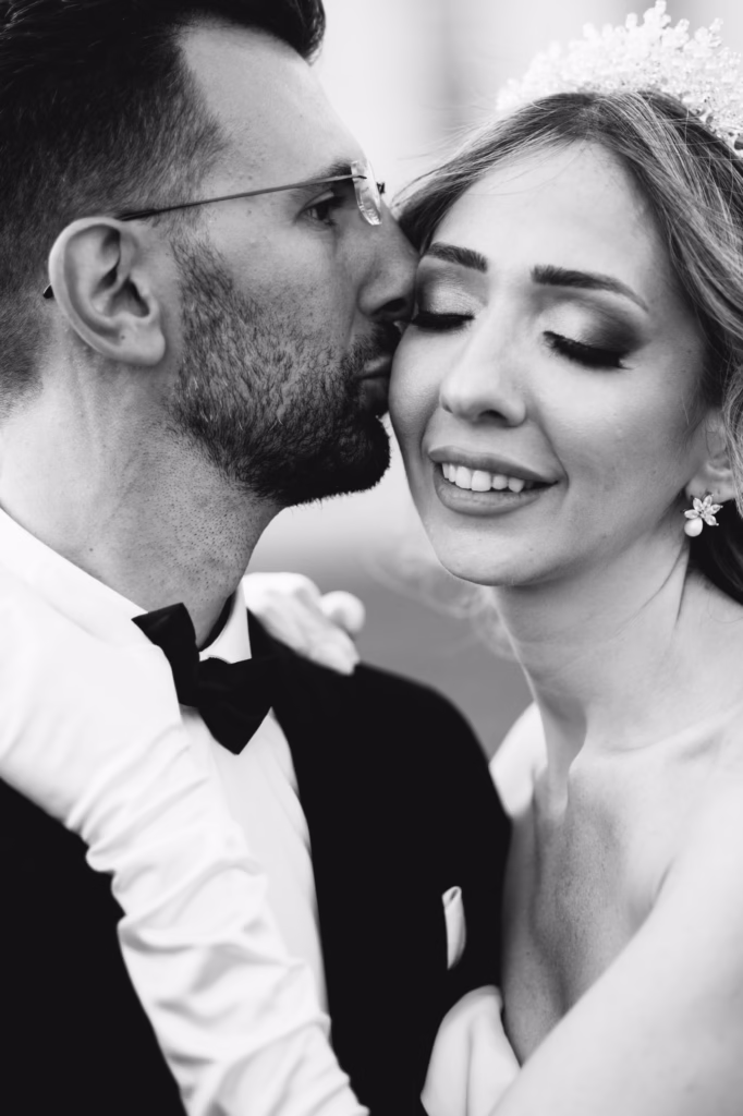 A bride and groom standing together in an intimate embrace, the groom gently kissing the bride’s cheek as she smiles softly, exuding romance and luxury.