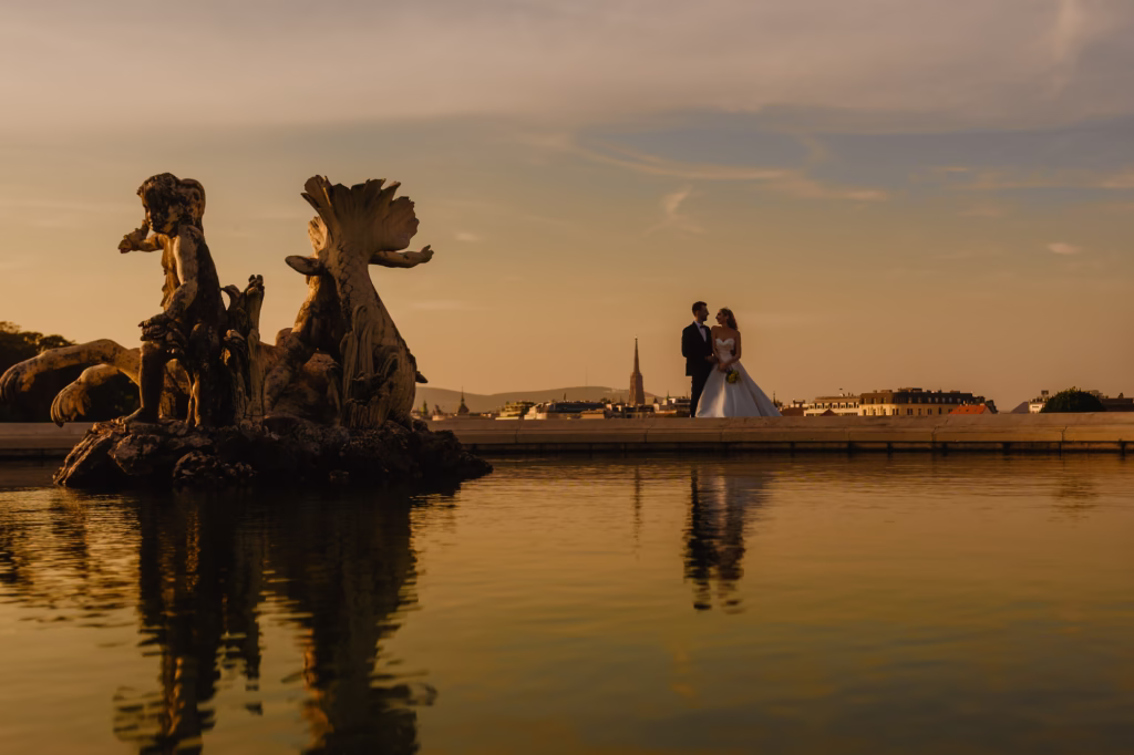 Bride and groom, Valentina & Valentin, stand near the grand fountain in Schönbrunn Palace Park, surrounded by lush gardens. The bride’s satin gown flows in the breeze, while the groom’s black tuxedo contrasts elegantly with the golden hues of the palace. A timeless, romantic wedding moment captured in Vienna.