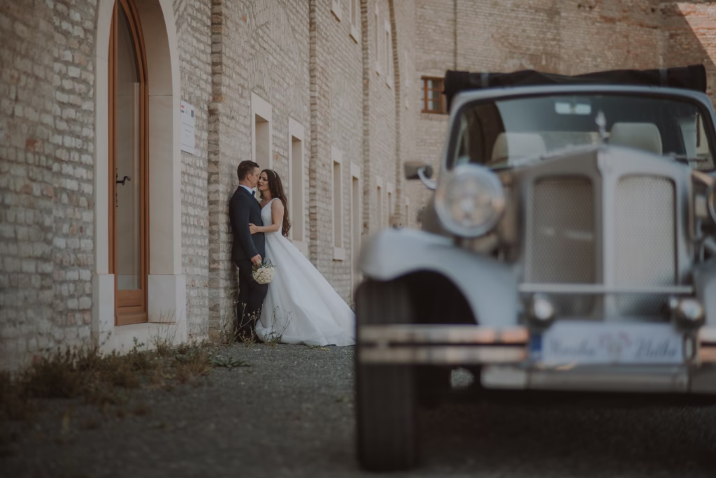 A bride and groom stepping into a vintage wedding car, her veil flowing gracefully as they prepare for their grand exit, framed by soft golden lighting.